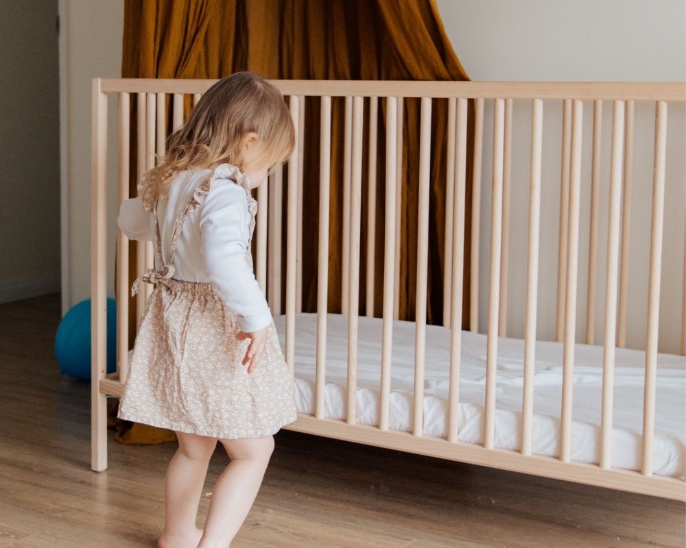 toddler girl standing next to crib on lowest setting