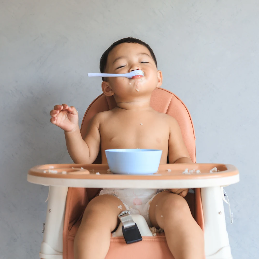 Baby boy sitting in a high chair, enjoying feeding himself rice. 