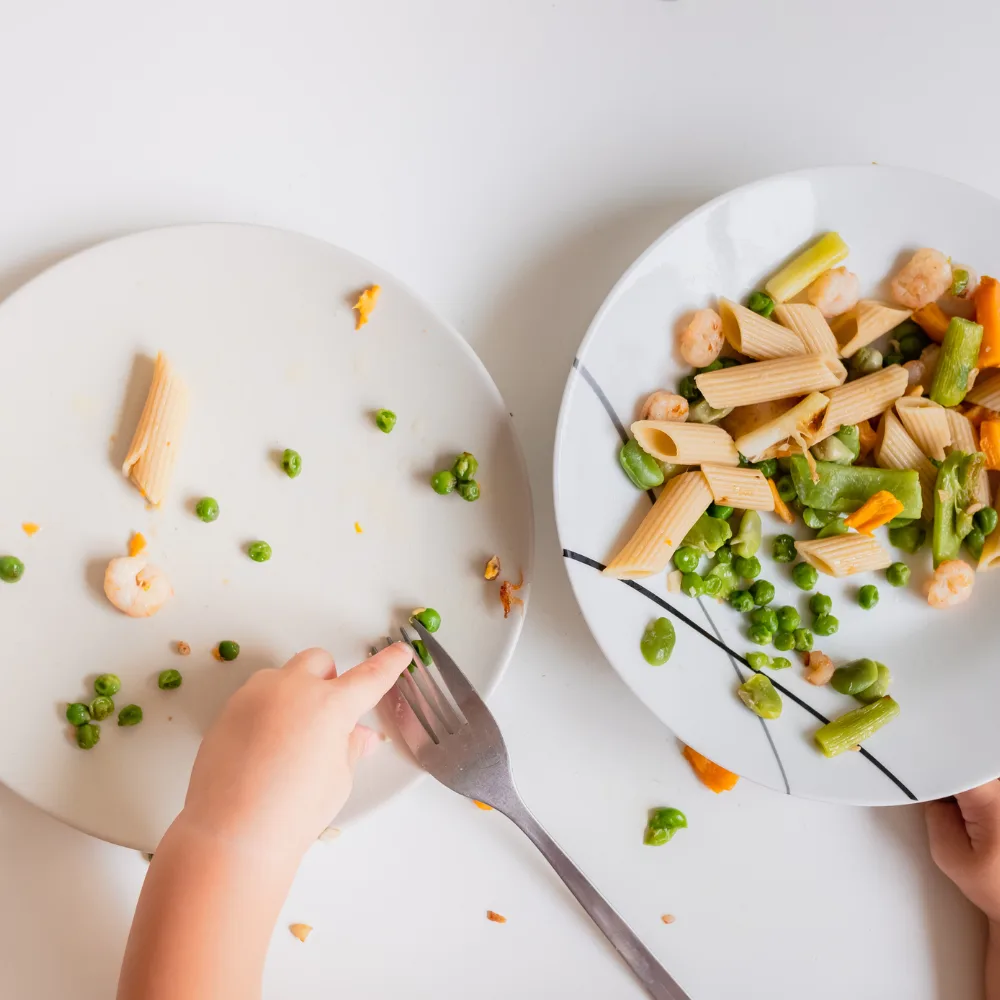 Two white plates with pasta, peas, and shrimp. A baby's hand is touching a metal fork sitting on the plate.