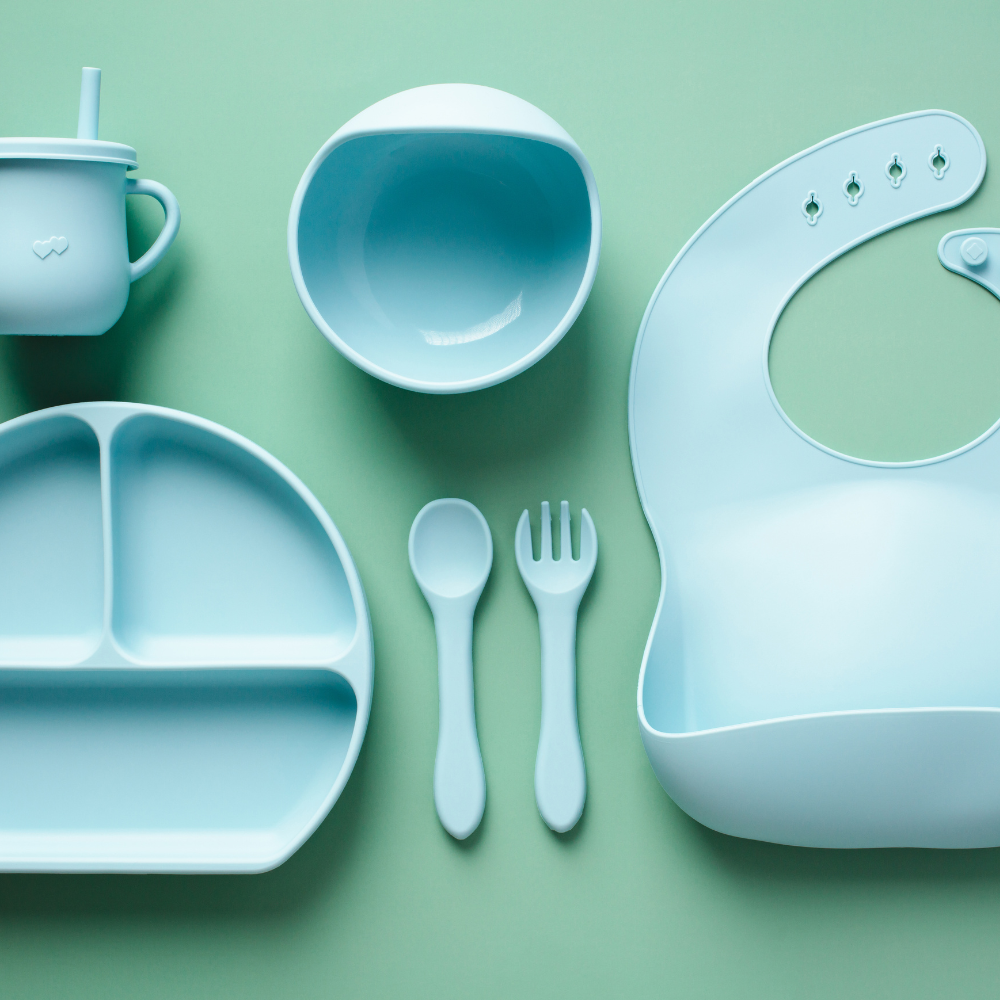 Silicone baby feeding supplies- cup, bowl, plate, bib, and fork and spoon layer out on a table.