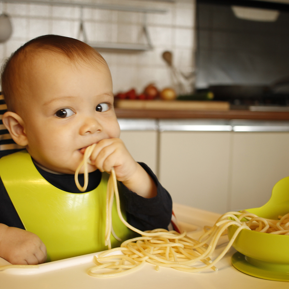 baby boy sitting in high chair, eating spaghetti noodles with his hand.