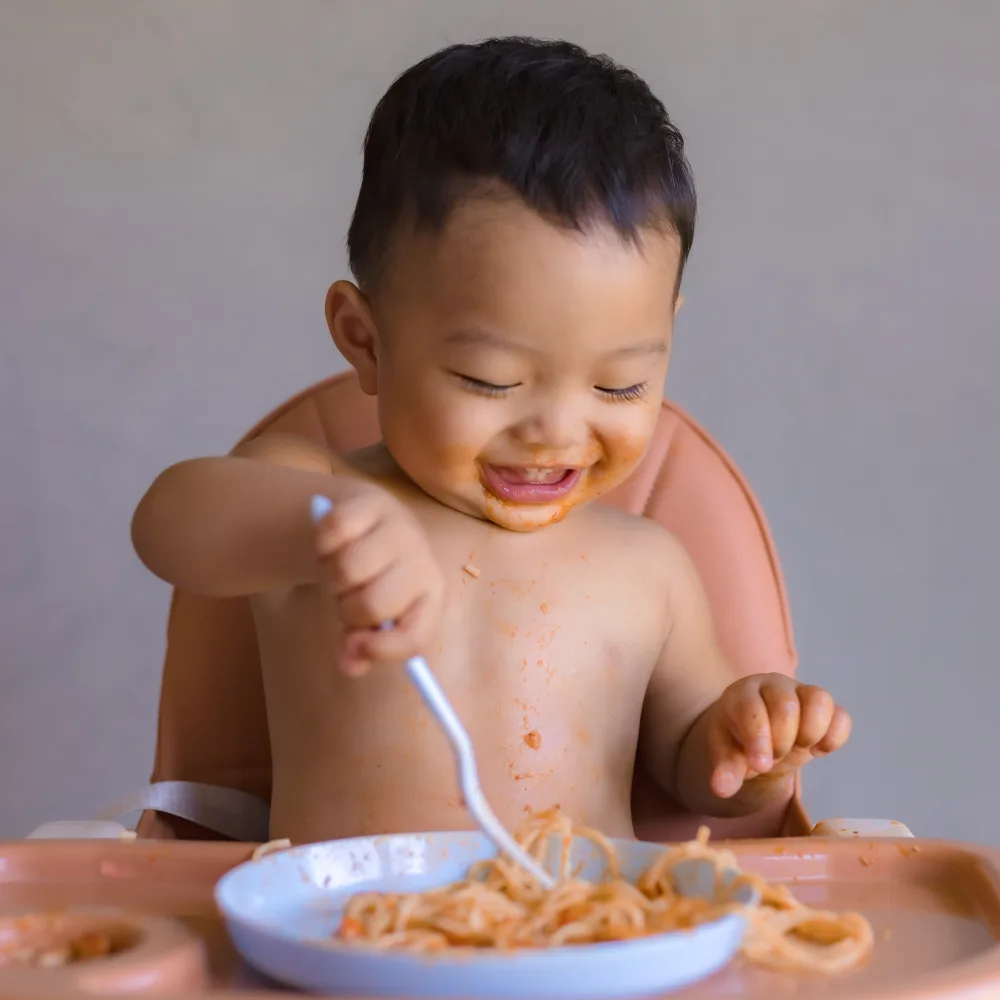 Baby with no shirt sitting in high chair, feeding himself spaghetti with sauce.