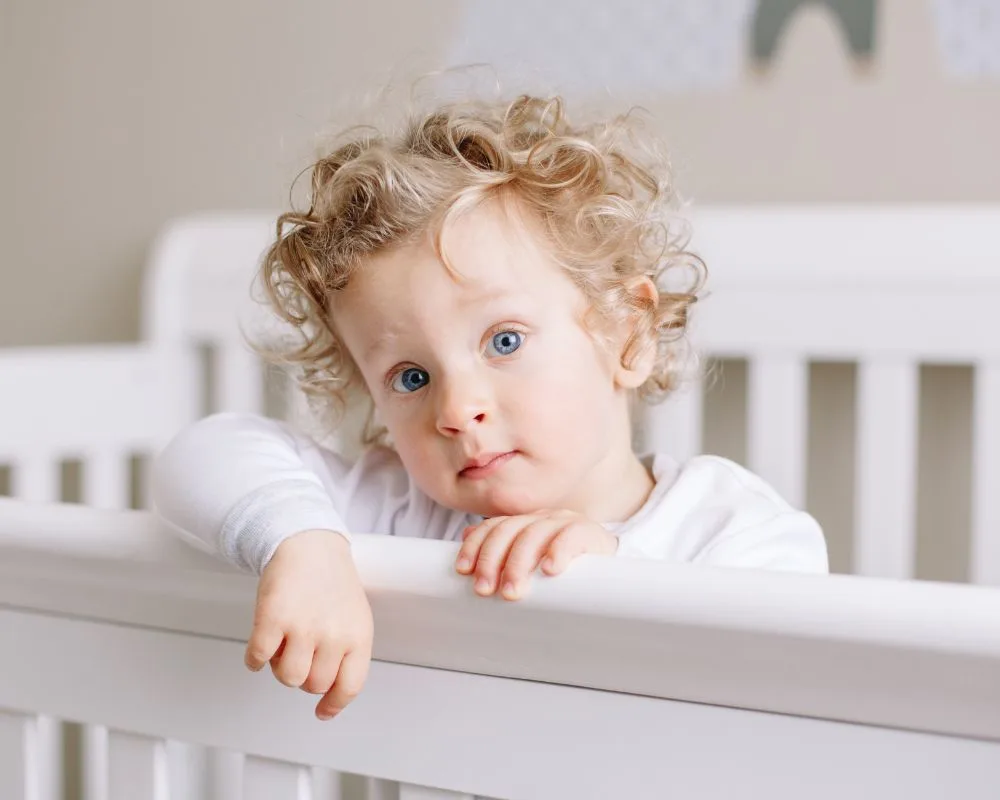 image of toddler standing inside crib with arm draped over the side