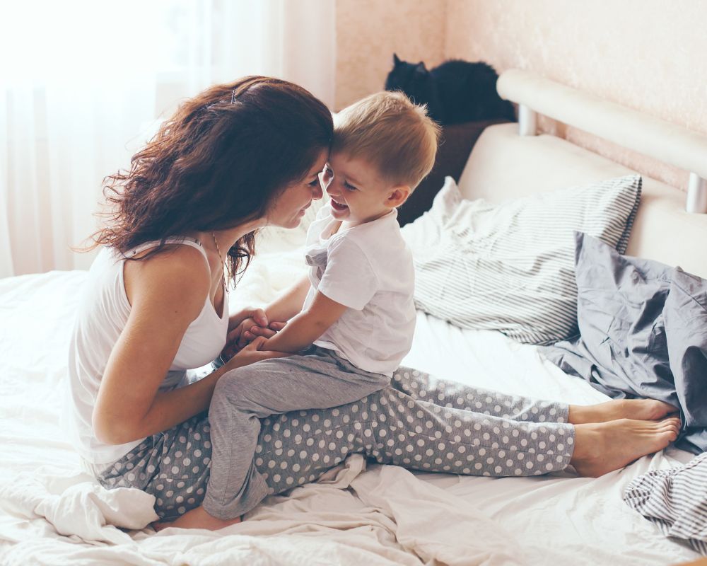 mom and toddler son laughing in bed in early morning